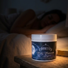 Jar of Aribella Moonlit Magnesium Cream on a bedside table with a person lying in bed in the background